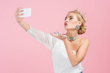 blonde woman in luxury crown taking selfie on smartphone and sending air kiss isolated on pink clipart