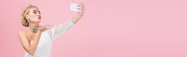 Lüks taçlı sarışın kadın akıllı telefondan selfie çekiyor ve pembe pankartta izole edilmiş bir şekilde hava öpücüğü gönderiyor.