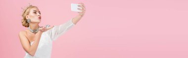 blonde woman in luxury crown taking selfie on smartphone and sending air kiss isolated on pink, banner clipart