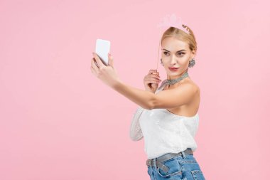 Çubukta kağıt taç tutan ve pembe renkte izole edilmiş akıllı telefondan selfie çeken sarışın kadın.