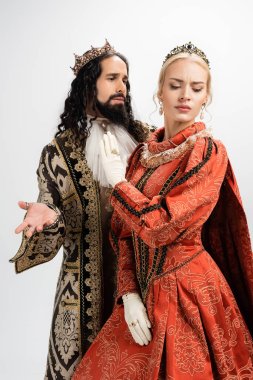 hispanic king in crown and medieval clothing looking at displeased blonde wife isolated on white