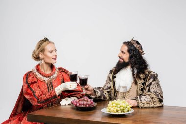 interracial historical couple in crowns clinking glasses of red wine while sitting at table with grapes isolated on white clipart