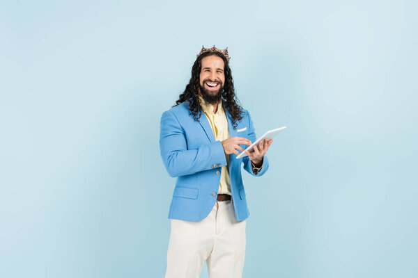 cheerful hispanic man in jacket and crown holding digital tablet isolated on blue
