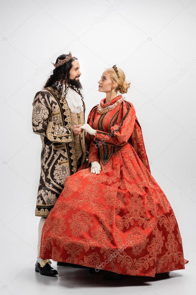full length of historical interracial couple in crowns and medieval clothing looking at each other and holding hands on white