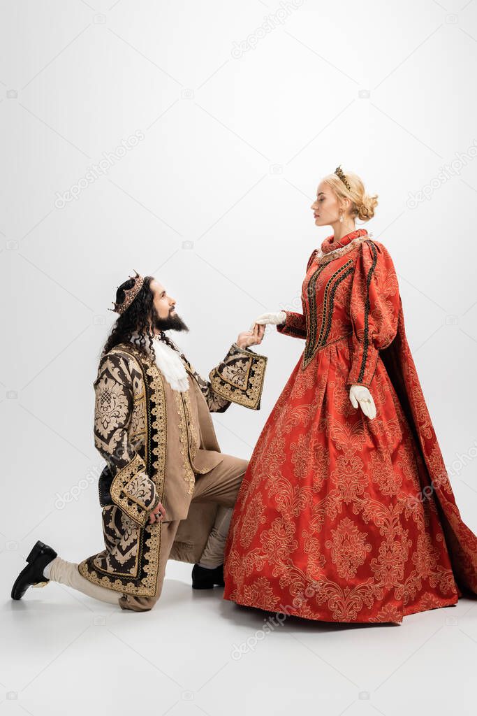 hispanic king in crown and medieval clothing standing on knee and holding hand of blonde queen on white