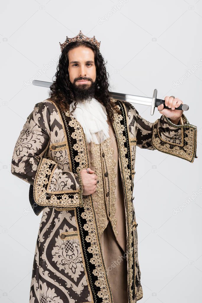 hispanic king in golden crown and medieval clothing holding metallic sword while looking at camera isolated on white