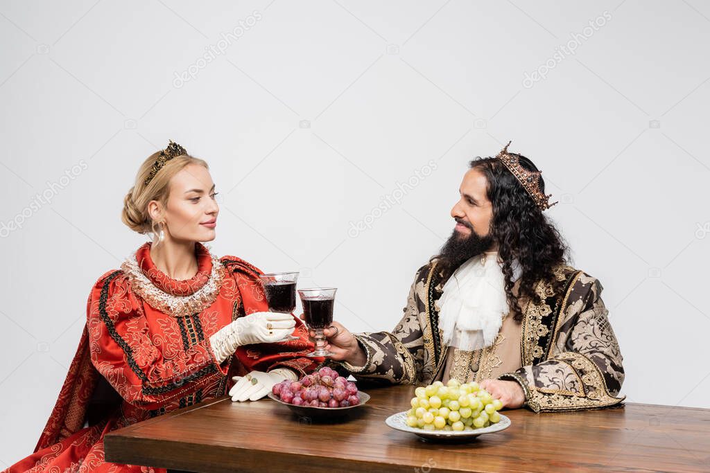 interracial historical couple in crowns clinking glasses of red wine while sitting at table with grapes isolated on white