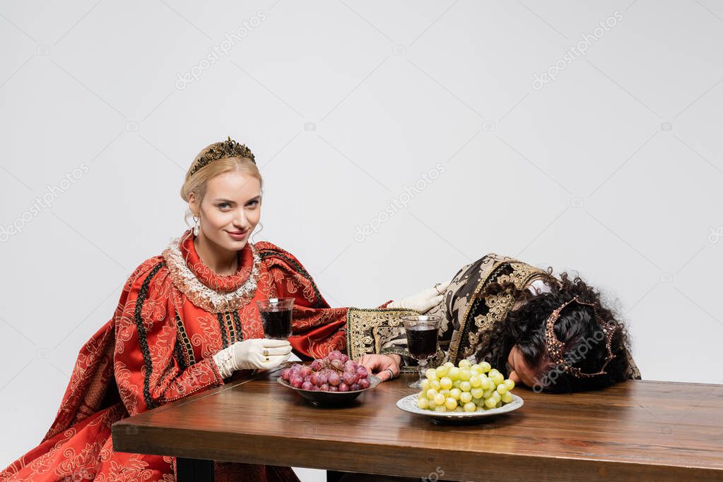 happy queen holding glass of red wine near poisoned hispanic king in medieval clothing isolated on white