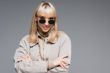 blonde and cheerful woman in sunglasses posing isolated on grey clipart