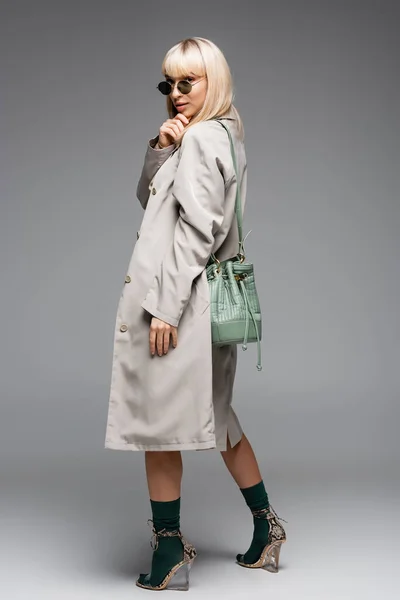 stock image full length of young woman in sunglasses and trench coat with green bag standing on grey