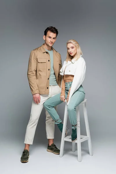 グレーの上に白い椅子の近くにポーズをとるパンツやジャケットのスタイリッシュなモデルの完全な長さ — ストック写真