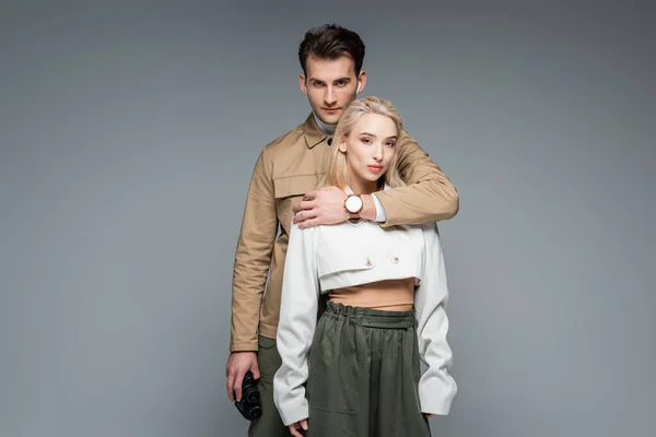 trendy man holding binoculars and hugging blonde woman isolated on grey