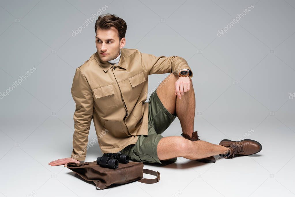 full length of trendy traveler in shorts and jacket sitting near leather backpack with binoculars on grey 