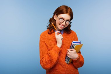 cheerful redhead student in glasses and orange sweater holding notebooks and pencil isolated on blue clipart