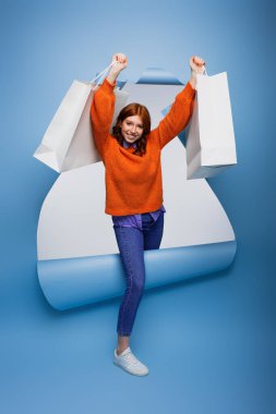 Mavi yırtık kağıt arka planda alışveriş torbaları tutan mutlu kızıl saçlı kadın. 