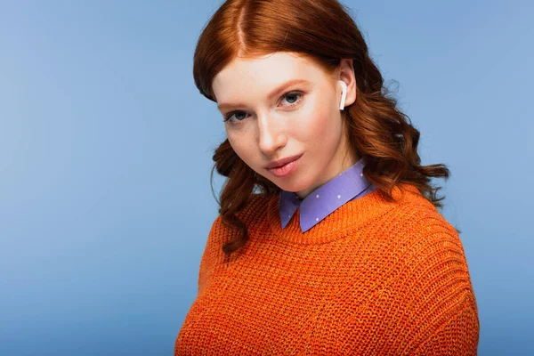 smiling redhead woman listening music in wireless earphones isolated on blue