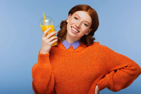 pleased young woman in sweater holding fresh orange juice in plastic cup isolated on blue