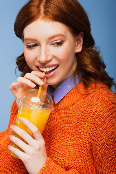 smiling redhead woman holding fresh orange juice in plastic cup isolated on blue