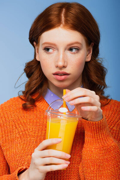 young redhead woman in sweater holding fresh plastic cup with orange juice isolated on blue