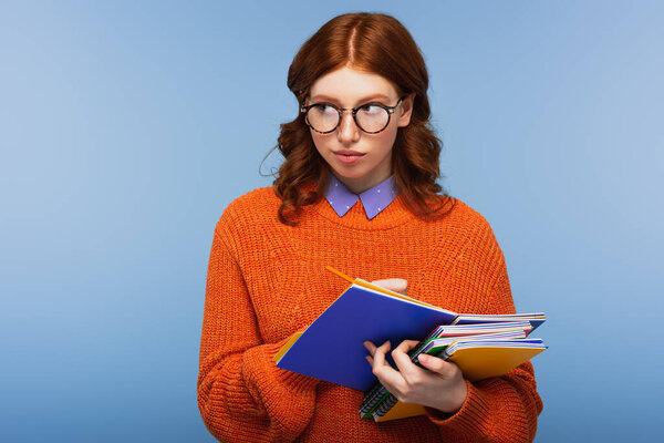 redhead student in glasses and orange sweater holding notebooks and pencil isolated on blue