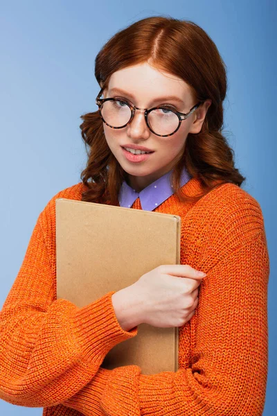 戴着眼镜 穿着橙色毛衣的红头发学生 手里拿着一本蓝色的书 — 图库照片