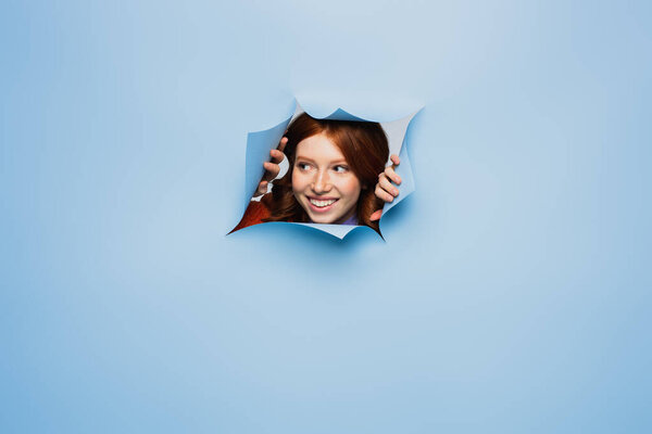счастливая рыжеволосая молодая женщина, отводящая взгляд на синем разорванном фоне