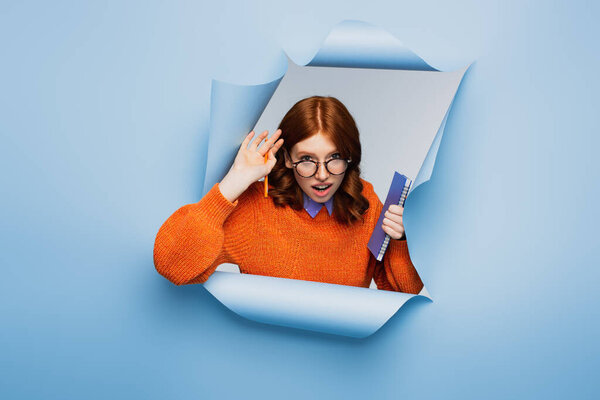 удивлена молодая женщина в оранжевом свитере и очках держа ноутбук и карандаш на синем разорванном фоне 