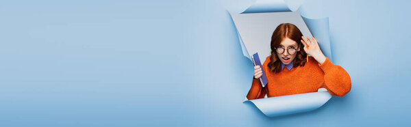 удивлена молодая женщина в оранжевом свитере и очках с блокнотом и карандашом на синем разорванном фоне, баннер