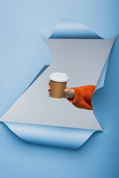 cropped view of young woman holding paper cup with coffee to go on blue ripped paper background 