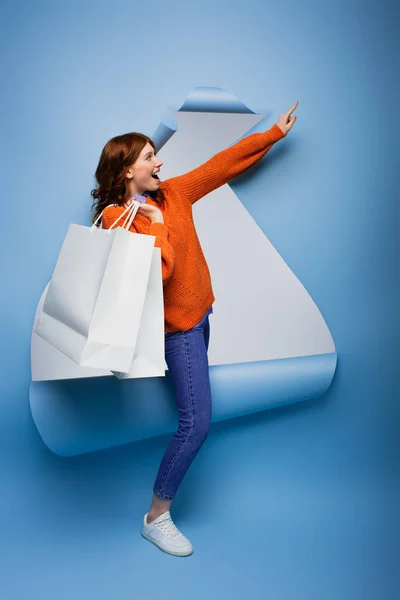 若いです驚くべき女性保持ショッピングバッグとジェスチャー上の青リッピング紙の背景 — ストック写真