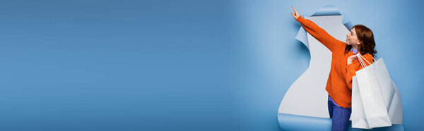 молодая счастливая женщина, держащая сумки с покупками и указывая пальцем на синий рваный бумажный фон, баннер