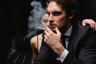 man in suit smoking cigarette near woman on blurred black background  clipart