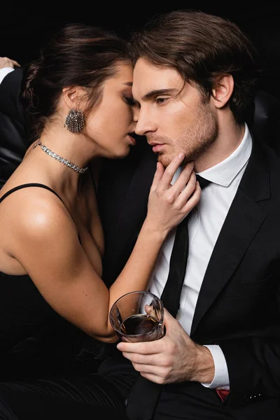 woman in slip dress seducing man in formal wear holding glass of whiskey on black