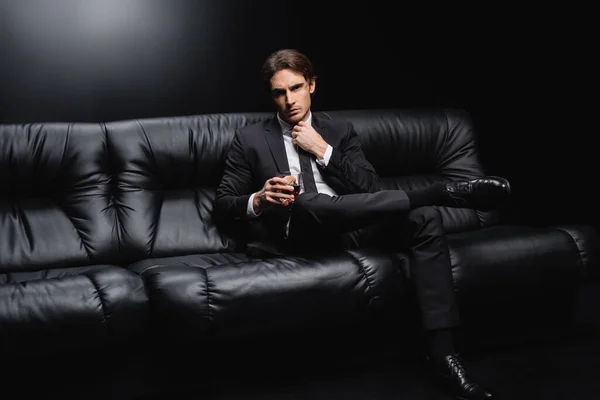 elegant man in suit adjusting tie and sitting on leather couch with glass of whiskey on black background