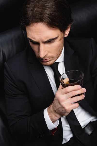 high angle view of elegant man in suit sitting on black leather couch with glass of whiskey