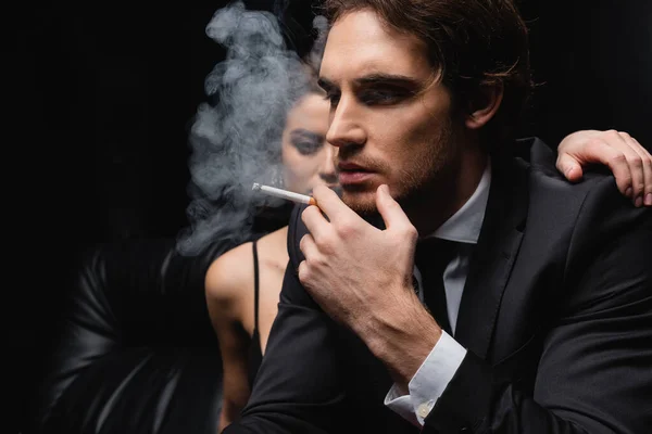man in suit smoking cigarette near woman on blurred black background