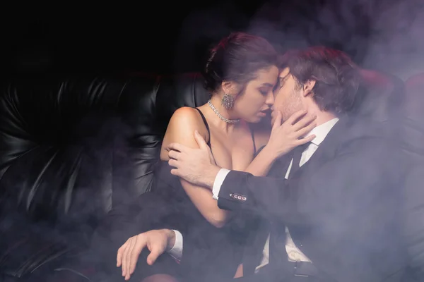 passionate woman in slip dress kissing man in suit on black with smoke