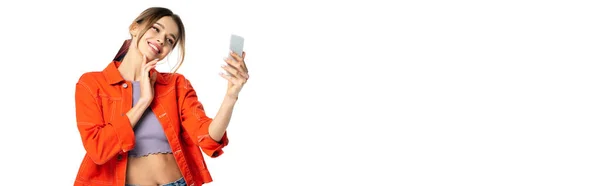 pleased young woman in crop top and orange shirt taking selfie on smartphone isolated on white, banner