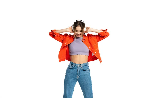 cheerful woman in jeans, crop top and orange jacket listening music in wireless headphones isolated on white