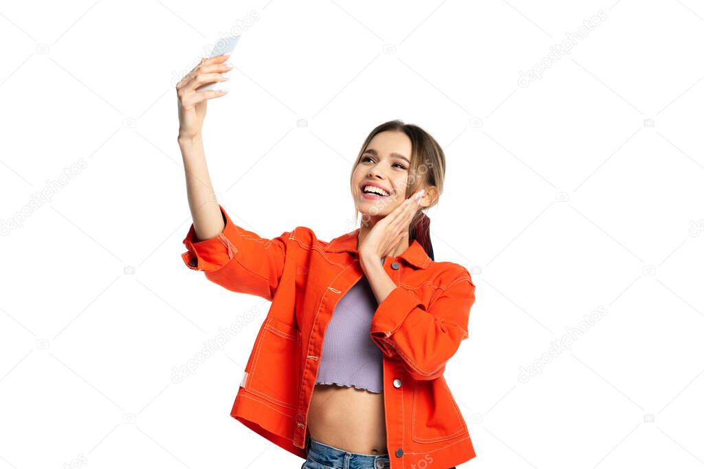 happy young woman in crop top and orange shirt taking selfie on smartphone isolated on white