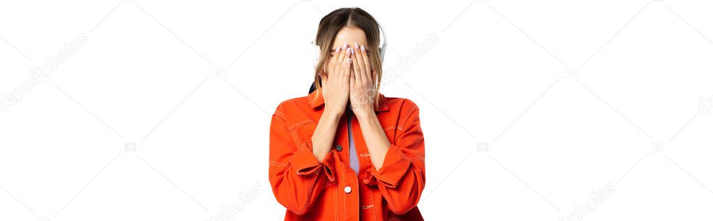 woman in orange shirt covering face while hands while listening music in wireless headphones isolated on white, banner