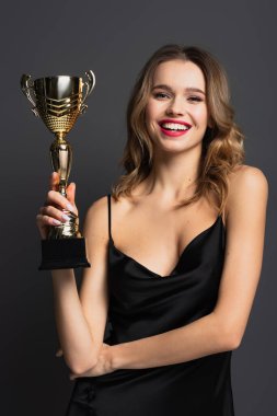 happy young woman in black slip dress holding golden trophy on grey clipart