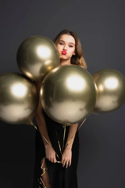 young woman in black slip dress holding golden balloons and pouting lips on grey