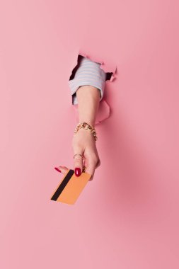 Cropped view of woman with red manicure holding credit card in hole of pink background  clipart