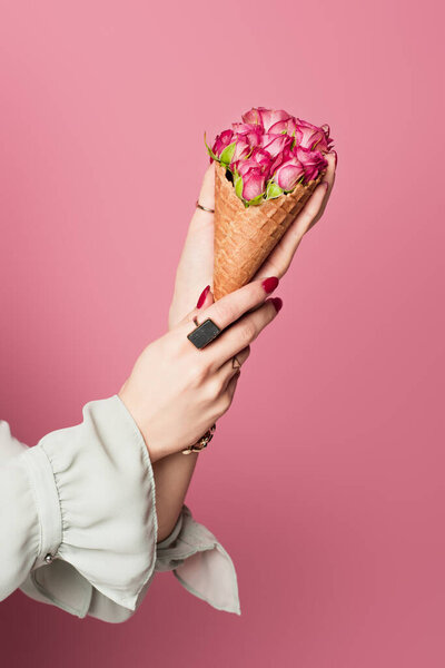 Cropped view of waffle cone with flowers in hands of woman isolated on pink 