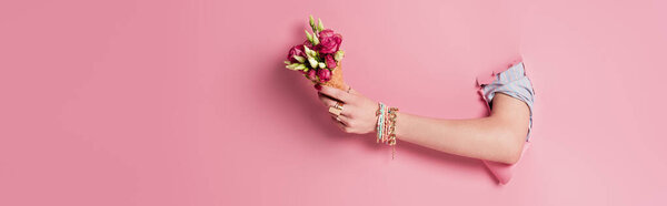Cropped view of woman holding delicious waffle cone and rose flowers in hole of pink background, banner