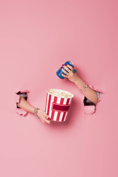 Cropped view of woman holding bucket of popcorn and drink near pink background with holes 