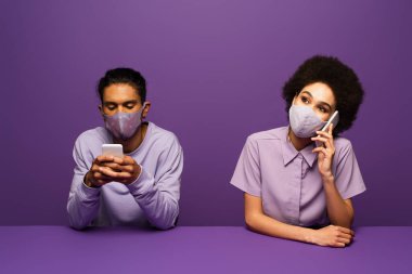 Mor üzerine izole edilmiş akıllı telefonlar kullanan koruyucu maskeli genç Afrikalı erkek ve kadın. 