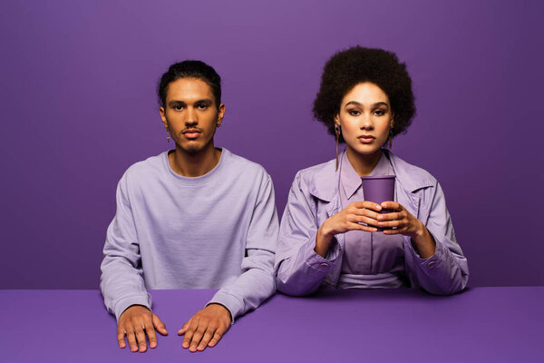 curly african american woman holding paper cup near man isolated on purple