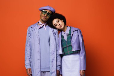 Mor ceketli kıvırcık kadın bereli modele yaslanıyor ve turuncu gözlükle izole edilmiş güneş gözlüğü var.